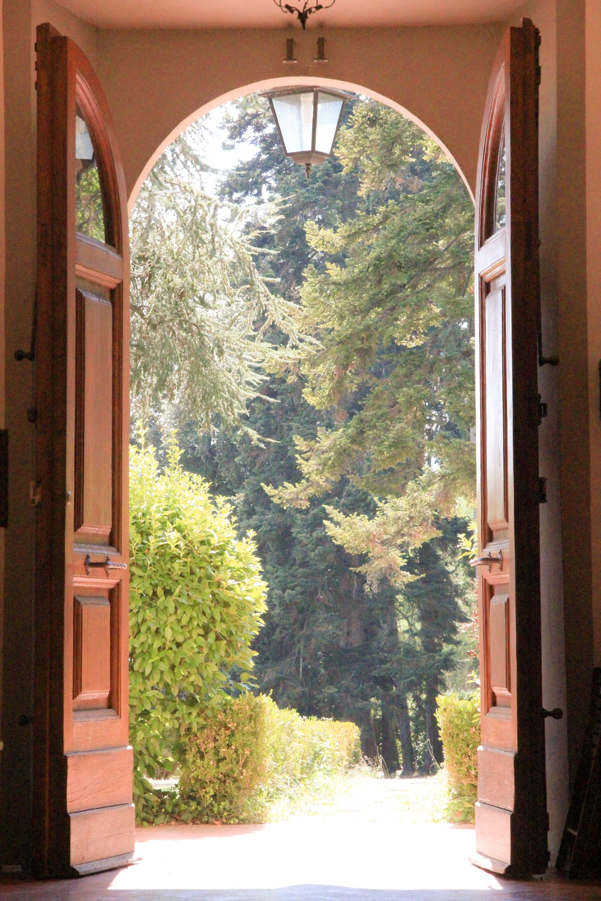 The entrance features tall, arched wooden doors that are widely opened. Sunlight illuminates the threshold, revealing a lush green garden framed by tall trees in the background, creating a serene connection to the outdoors.