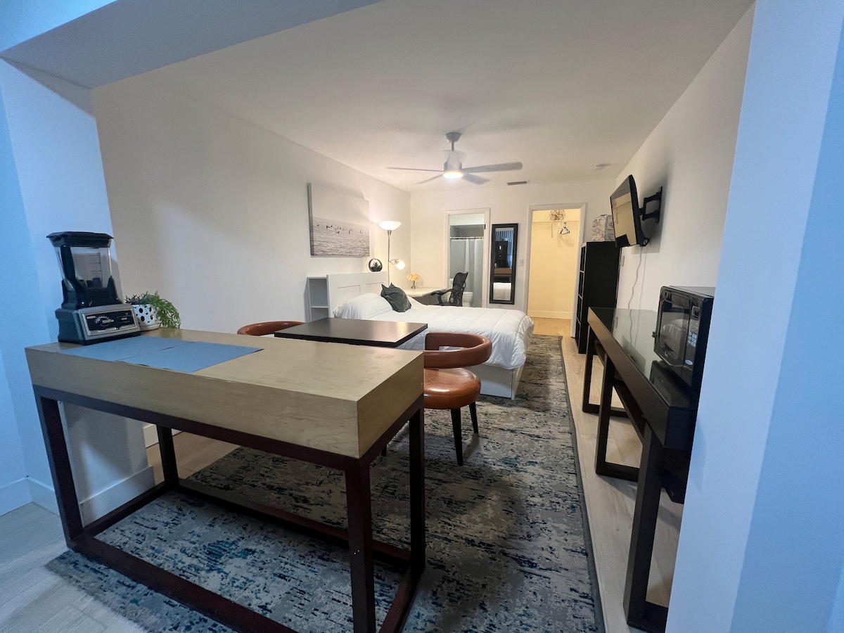 A spacious studio is presented, featuring a large bed and a functional workspace with a wood desk. A kitchenette area with a microwave and coffee maker is visible. Light-colored walls and a neutral rug add to the room's open feel. Additional furnishings include a flat-screen TV and a small table.