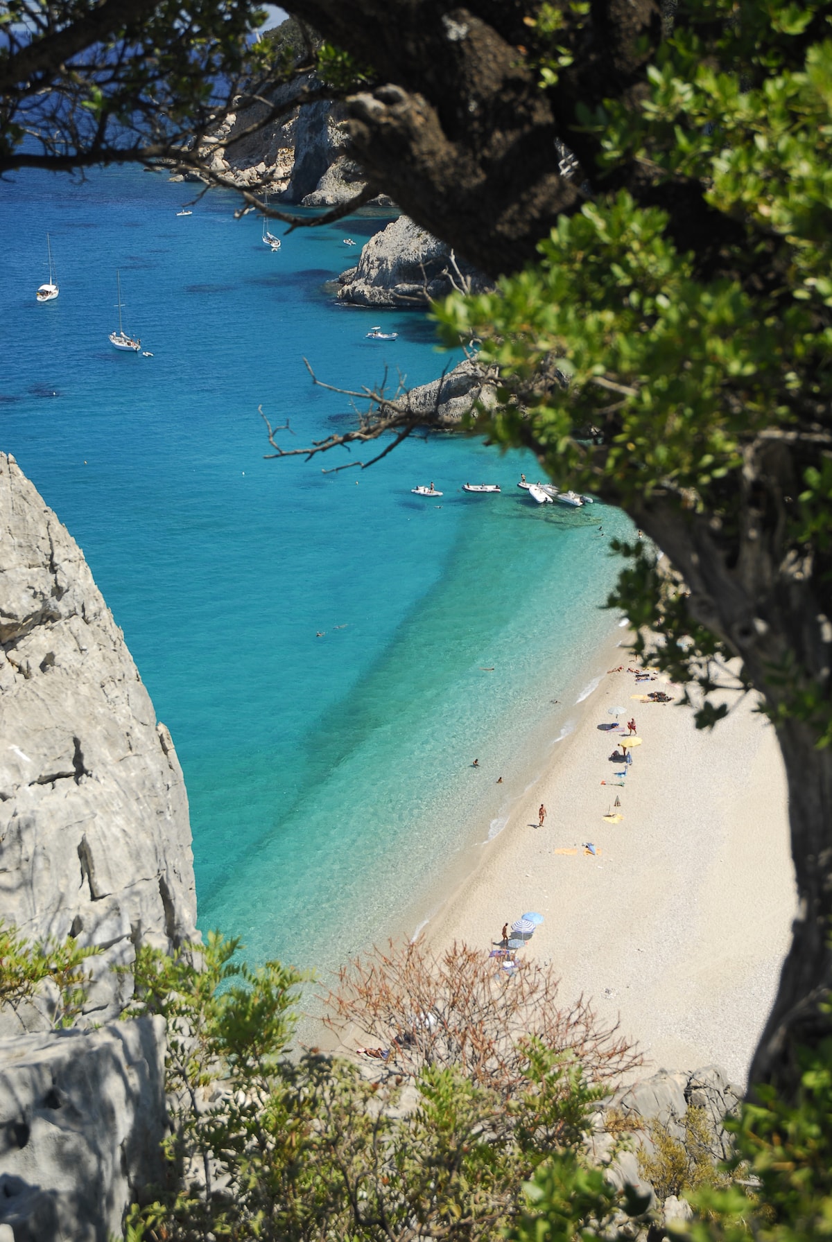 A serene beach scene is visible, framed by greenery and rocky cliffs. Clear turquoise waters gently lap against a sandy shore, where a few sunbathers enjoy the sun. Small boats are anchored nearby, contributing to the tranquil coastal atmosphere.
