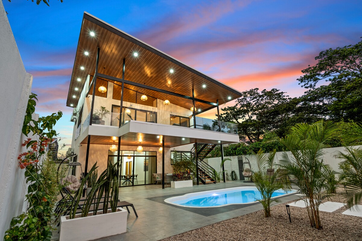 The two-story duplex apartment is framed by lush greenery and features a private pool surrounded by a spacious outdoor area. The modern structure is adorned with illuminated lanterns and expansive glass doors, offering views of the vibrant sky at sunset.