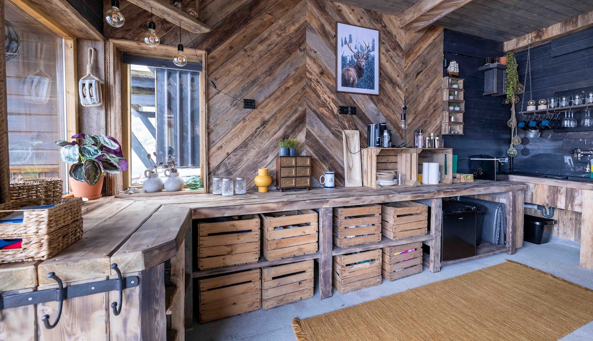 A rustic kitchen features wooden cabinetry and shelving with various utensils and glassware visible. The countertop is adorned with potted plants and decorative items. Below, wooden crates serve as storage, while a cozy rug lies on the floor, adding warmth to the space.