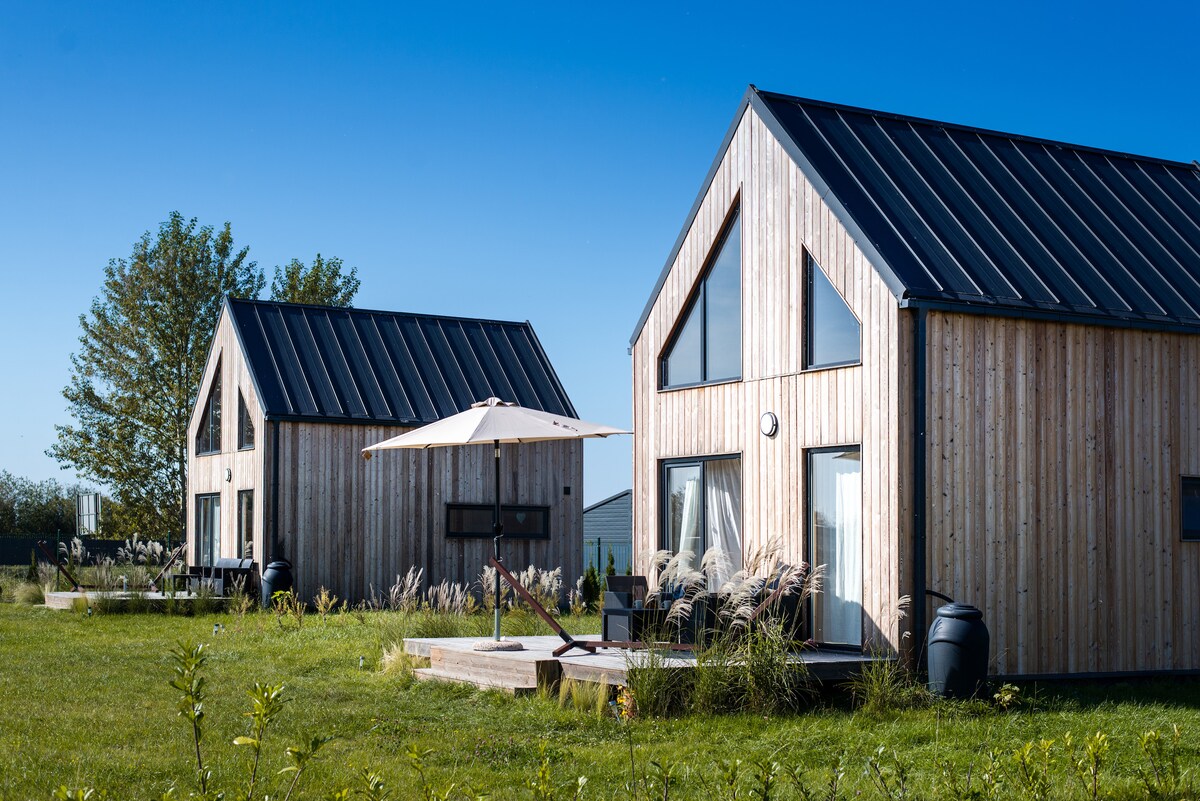 Two wooden cottages are set in a spacious green area under a clear blue sky. Each cottage features large windows and a sloped roof with a dark finish. A shaded patio area is visible, complete with an umbrella and outdoor seating.