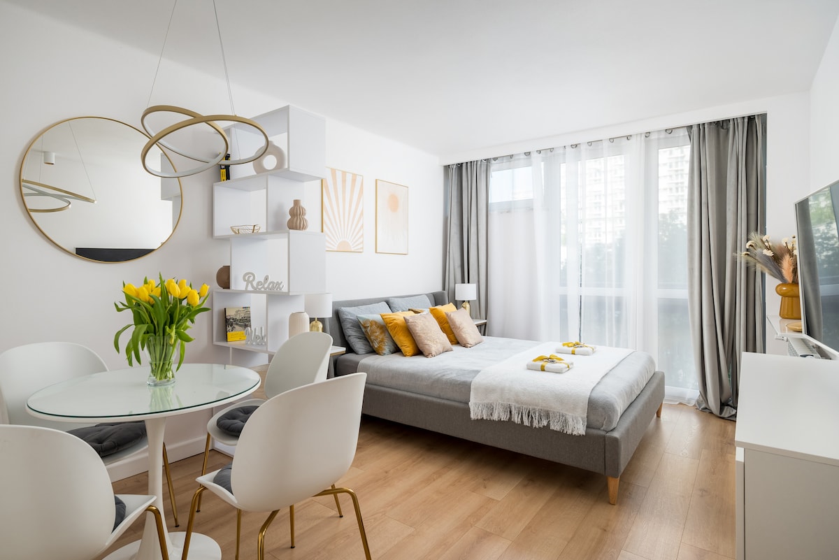 A bright living area features a spacious bed adorned with colorful pillows and a light throw. A circular mirror and modern pendant light are showcased above a glass dining table, surrounded by white chairs. Large windows allow natural light to fill the room.