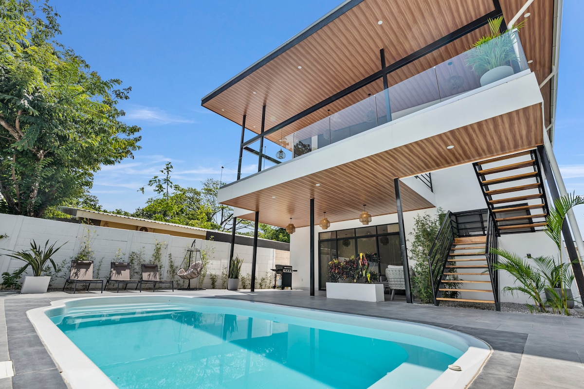 A modern villa featuring a private swimming pool surrounded by a sleek deck. The upper level showcases expansive glass railings and a wooden overhang, with ample outdoor seating. Lush greenery and tropical plants enhance the serene atmosphere, while a staircase provides access to the upper floor.
