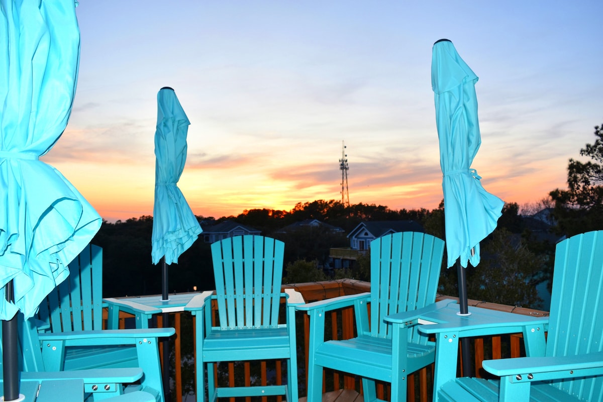 A treetop deck is highlighted with bright turquoise outdoor chairs, each paired with a matching umbrella. The sky transitions to vibrant hues of orange and purple at sunset, providing a scenic backdrop to the tranquil outdoor seating area.