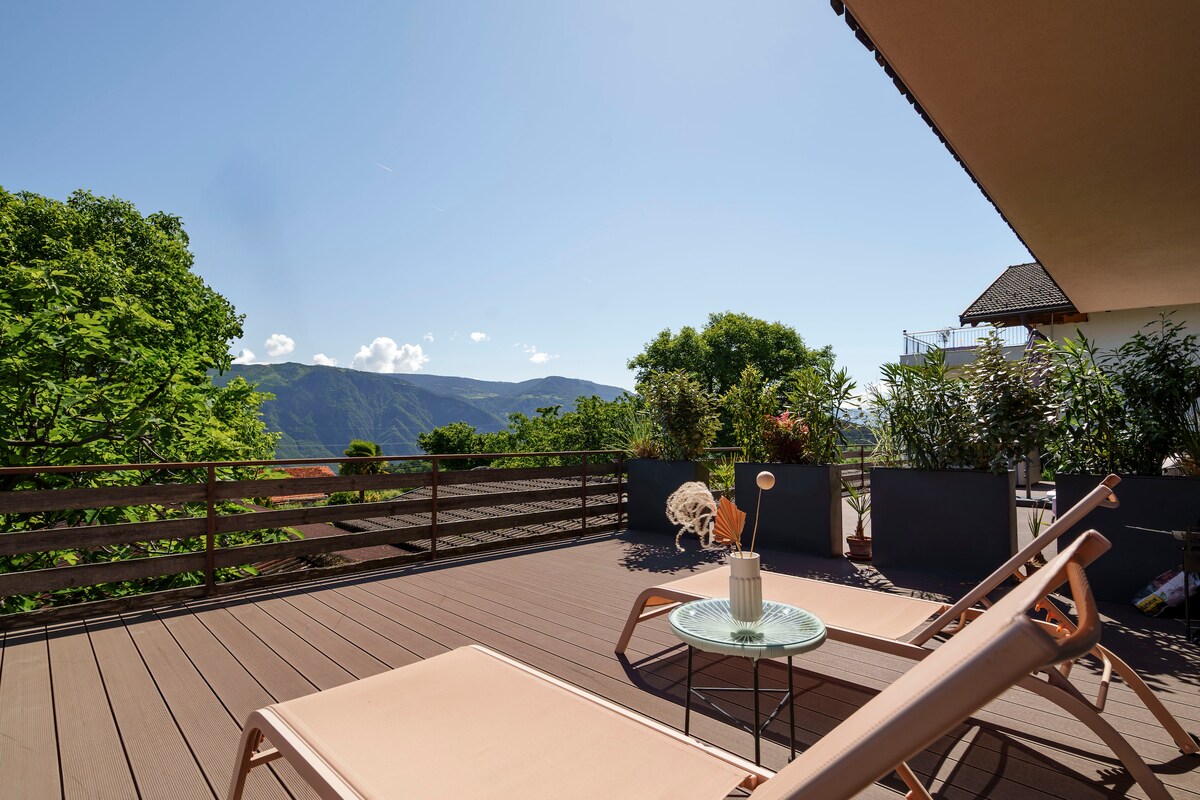 A spacious terrace is highlighted by two lounge chairs, providing a view of the surrounding mountains and greenery. Potted plants add a natural touch, while a small table sits between the chairs, enhancing the inviting outdoor space.