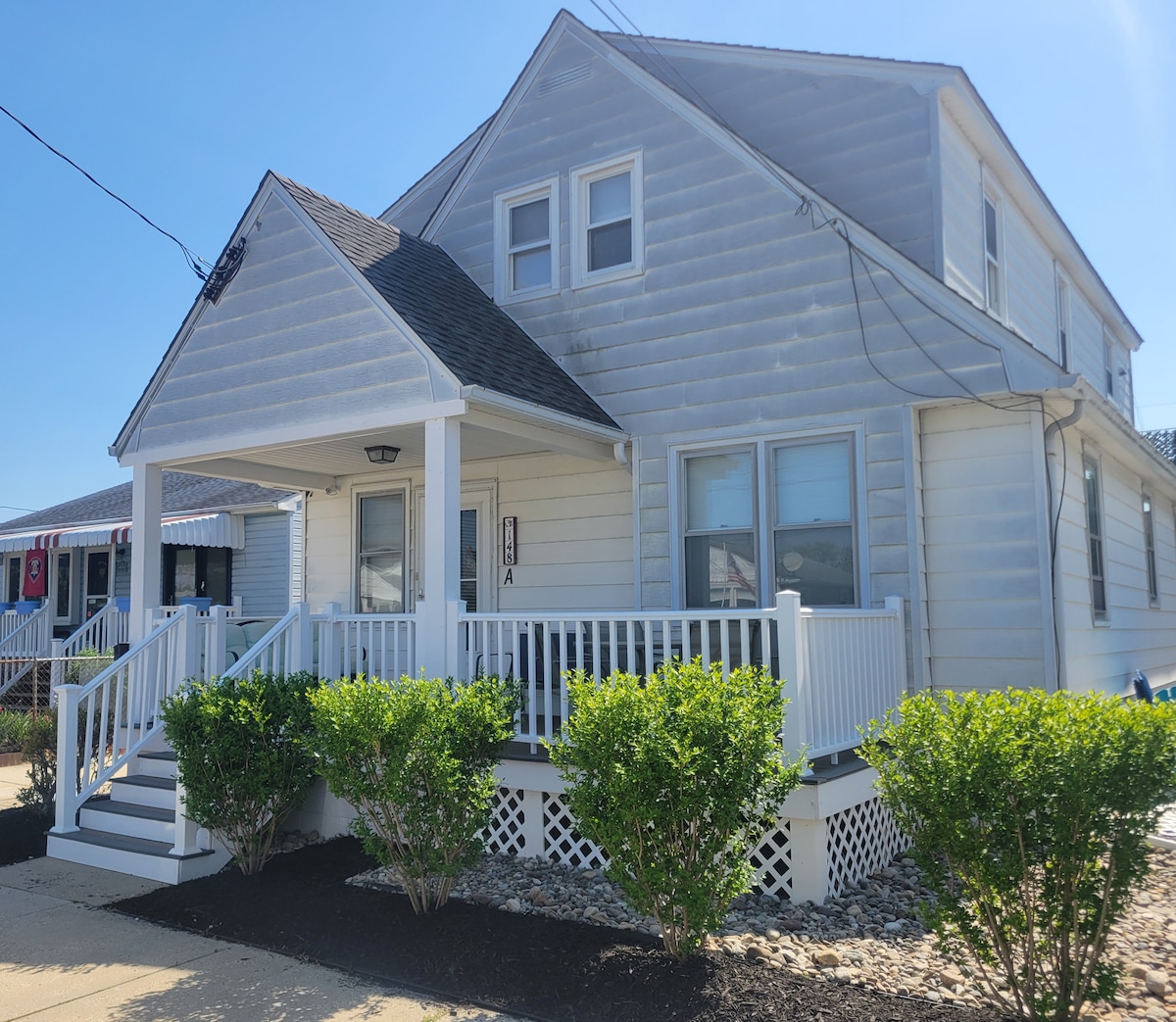 A well-maintained exterior of a two-story building is showcased, featuring a covered porch with white railings and stairs leading to the entrance. Green shrubs line the front, adding to the welcoming look of the property under clear blue skies.