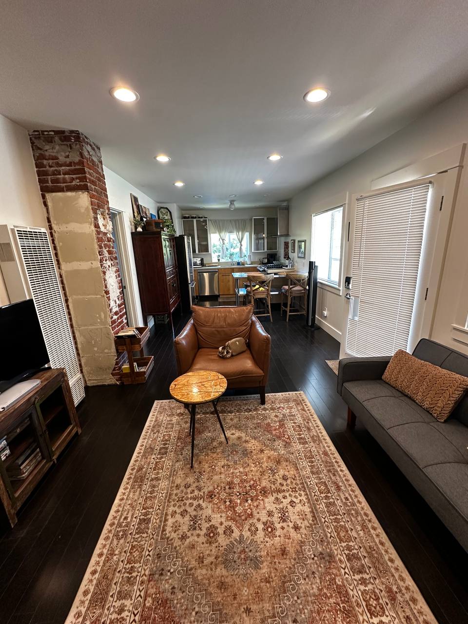 A cozy living area is equipped with a leather armchair and a grey sofa, centered around a small circular table. A decorative area rug adds warmth to the dark hardwood floor. Bright lighting illuminates the space, leading to a view of the kitchen through an open layout.