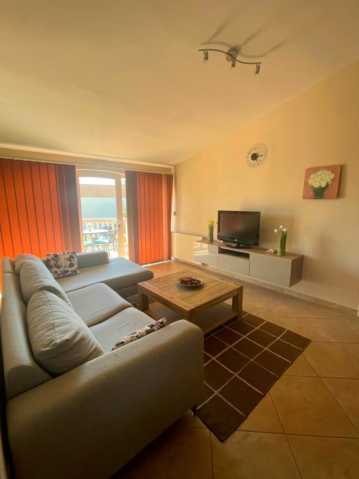 A bright living area features a comfortable gray sofa and a wooden coffee table, adorned with a decorative plate. Large glass doors provide access to a terrace, while vertical orange blinds filter in natural light. A television is positioned on a sleek white cabinet.
