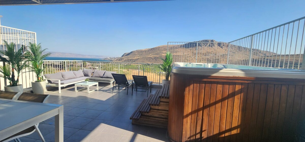A spacious balcony features comfortable seating with sofas and sun loungers. A hot tub is visible, surrounded by wooden accents. The backdrop showcases a scenic view of hills and a clear sky, adding to the expansive outdoor space.