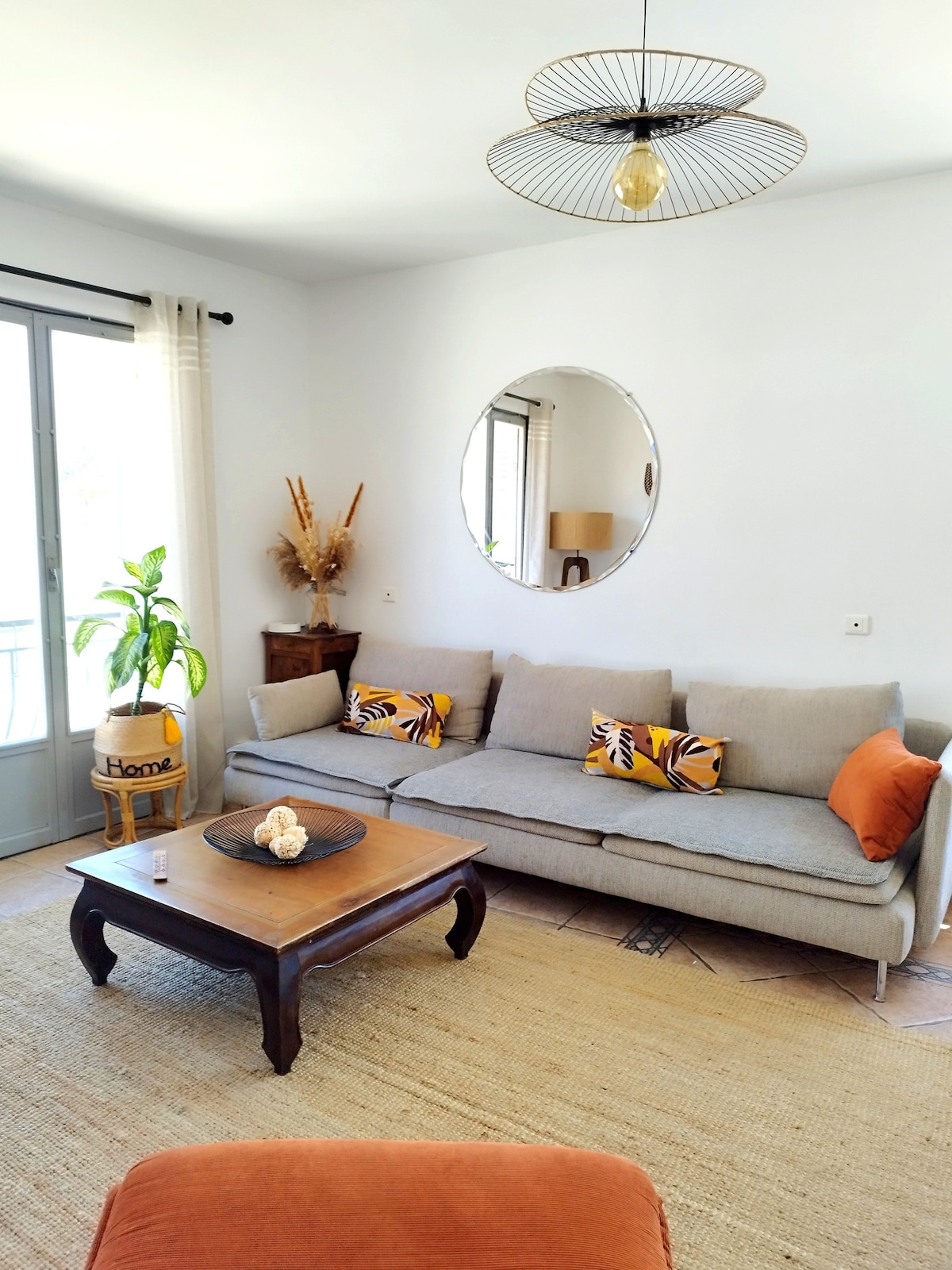 A comfortable living area showcases a light gray sofa adorned with patterned cushions and an orange accent pillow. A round mirror reflects light, while a wooden coffee table sits at the center on a woven rug, enhancing the serene atmosphere. A potted plant adds a touch of greenery.
