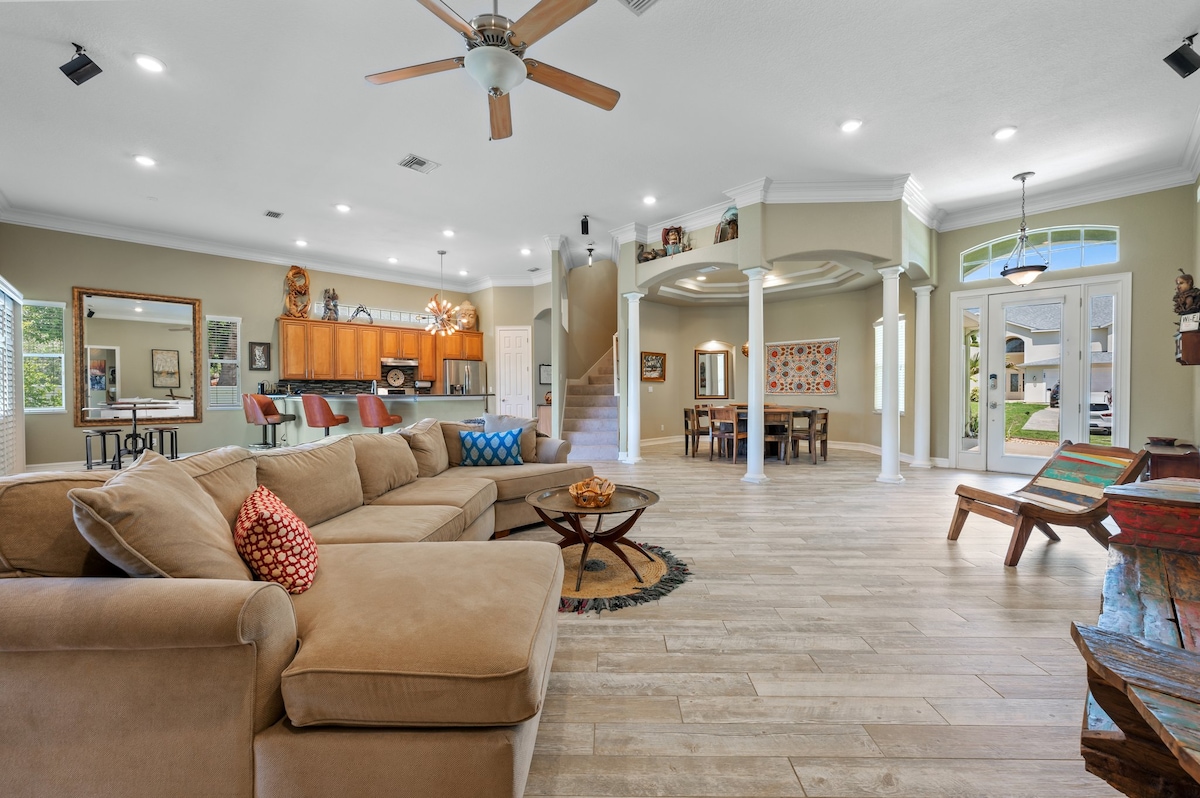 A spacious great room features a large sectional sofa with decorative pillows, inviting relaxation. The open layout connects seamlessly to the modern kitchen and dining area, which are enhanced by natural light from multiple windows. Decorative artwork and colorful furnishings are showcased throughout.