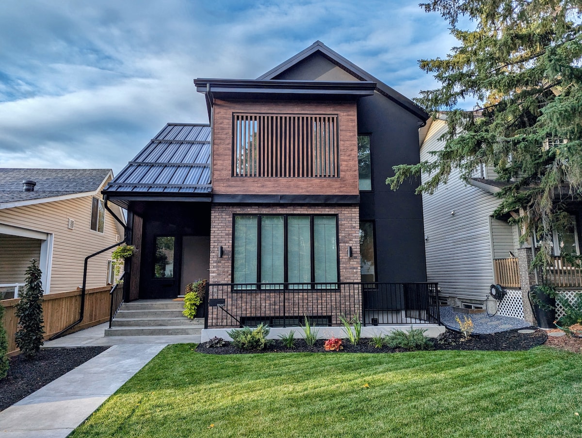 A modern two-story home features a combination of dark siding and brick with a geometric design. Large windows provide natural light, while a well-maintained lawn with various plants enhances the exterior. A paved pathway leads to the entrance, adding to the inviting aspect of the property.