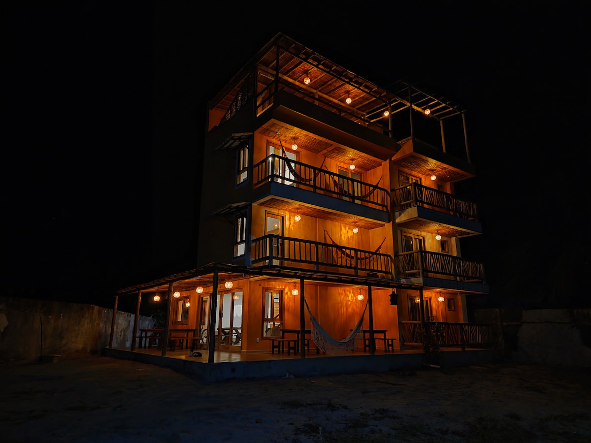 The three-story house is illuminated at night, highlighting its balconies and warm light emanating from the windows. The structure features a spacious patio area with tables, offering a relaxed setting surrounded by nature.