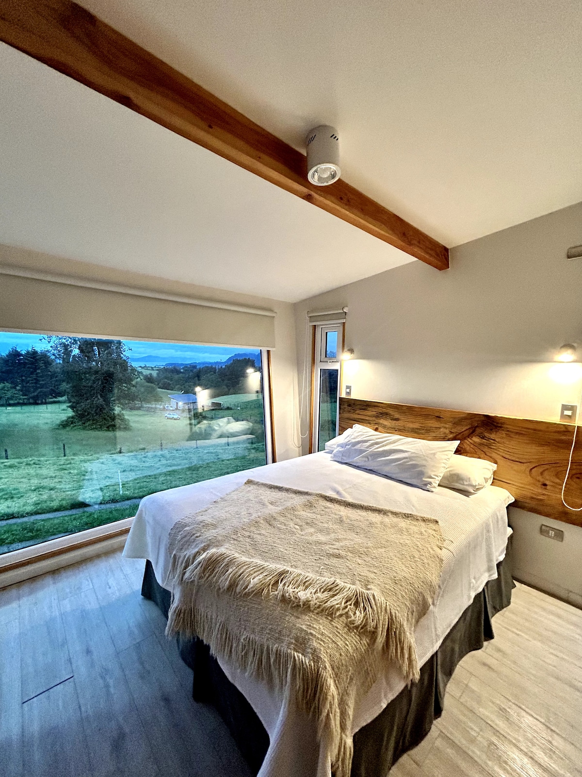 A well-appointed sleeping area features a spacious bed with a soft blanket draped across it. Large windows allow natural light and offer views of the surrounding fields and hills, while wooden beams add a touch of warmth to the room.