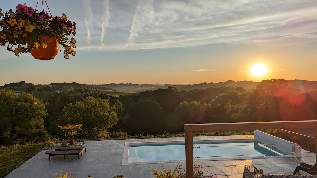 A serene sunrise casts a warm glow over the landscape, with a clear view of rolling hills and lush greenery. A swimming pool is visible in the foreground, complemented by comfortable lounge chairs. Vibrant flowers hanging above add a touch of color.