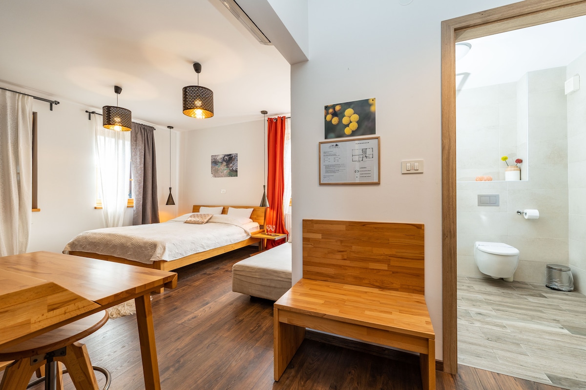 A spacious room features a comfortable bed with soft linen and warm lighting from stylish hanging fixtures. A wooden dining table and benches complement the neutral-toned decor. An en-suite bathroom is visible, showcasing modern fixtures and natural light.