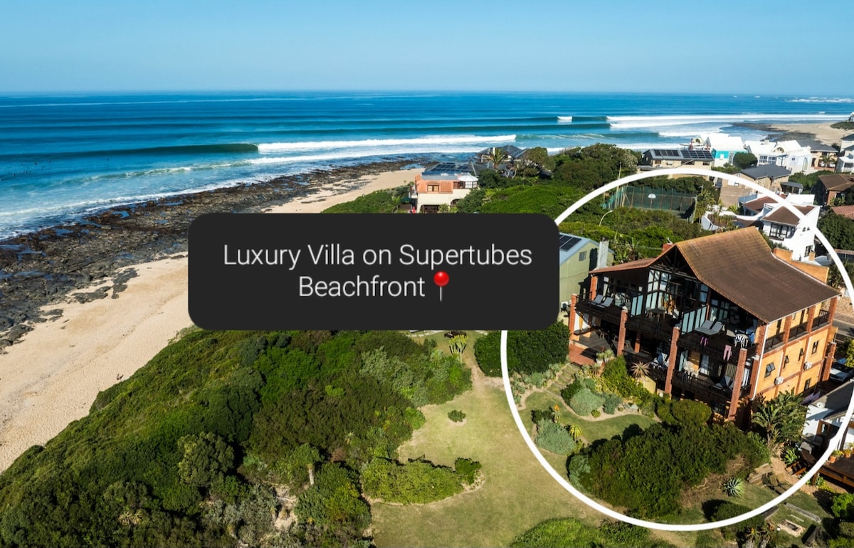 An aerial view showcases a luxury villa situated on the Supertubes beachfront. Sandy shores and rolling waves are visible, with the villa surrounded by lush greenery. The image captures the proximity to the ocean, enhancing the serene coastal setting.
