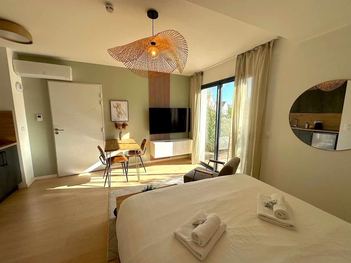 A spacious living area features a large bed with neatly arranged towels on top. A flat-screen TV is mounted on the wall, and natural light floods through a sliding glass door leading to the outdoors. A dining table and chairs are positioned near the kitchenette.