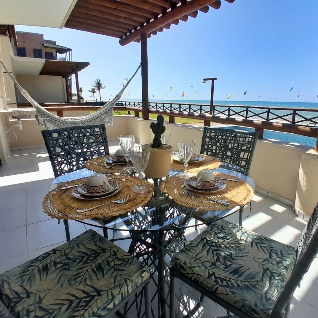 An outdoor dining area is shown, featuring a glass table with four chairs, each adorned with a leafy design. A simple centerpiece with a small plant is present. A hammock is seen in the background, with a view of the ocean and colorful kites visible in the distance.