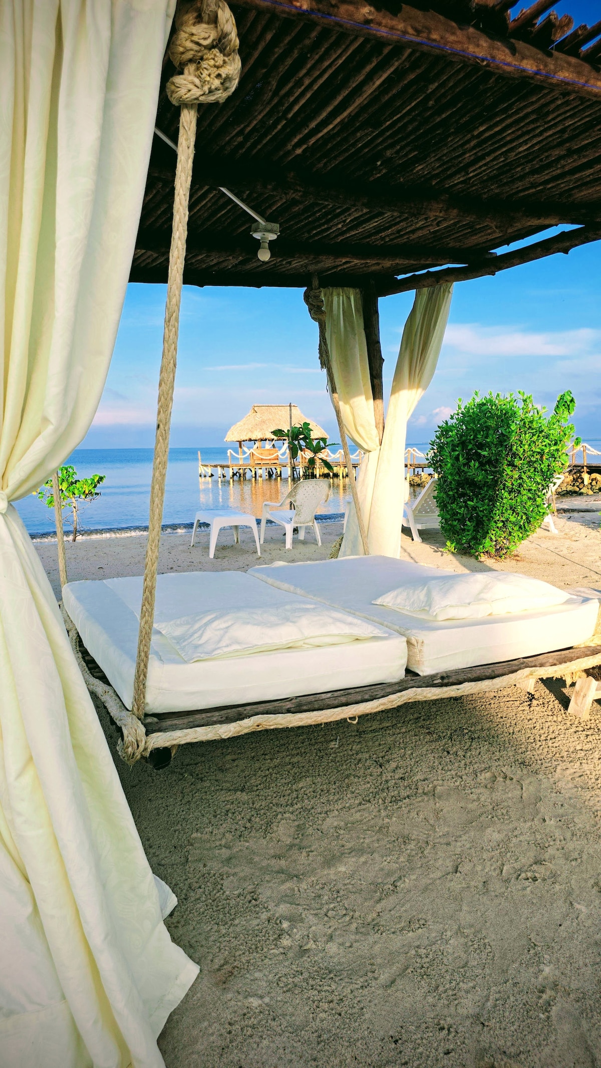 A shaded swing bed, draped with light fabric, provides a relaxing space overlooking the sandy beach. Soft hues from the setting sun illuminate the tranquil waters in the distance, while small white tables and chairs can be seen nearby, enhancing the serene coastal atmosphere.