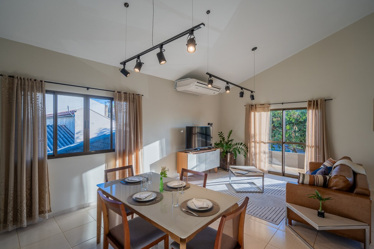 A well-lit living area features a modern dining table set for four, accompanied by a comfortable seating arrangement. Large windows allow natural light to illuminate the space, while indoor plants add a touch of greenery. A smart TV is positioned against a white media console.