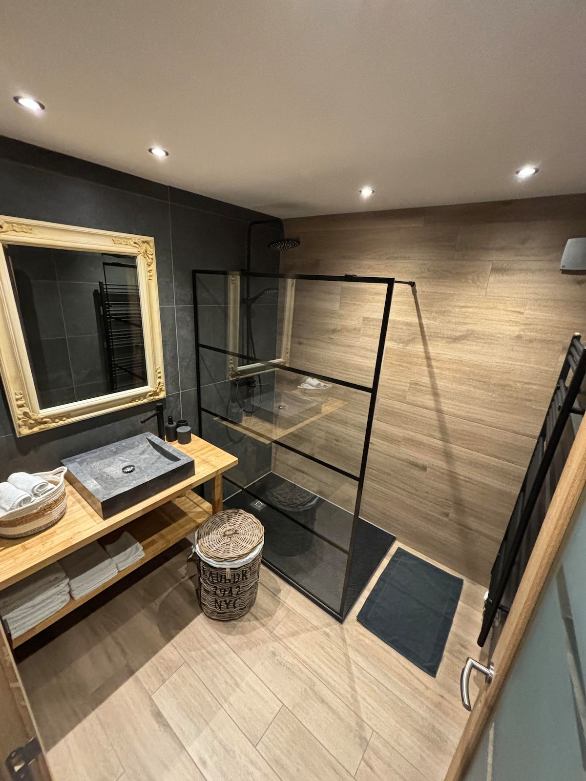 The bathroom features a glass-enclosed shower with modern fixtures, complemented by wooden accents. A large mirror is framed elegantly above a wooden vanity with a stone sink. Fresh towels are neatly arranged on shelves, and a stylish basket adds a functional touch.