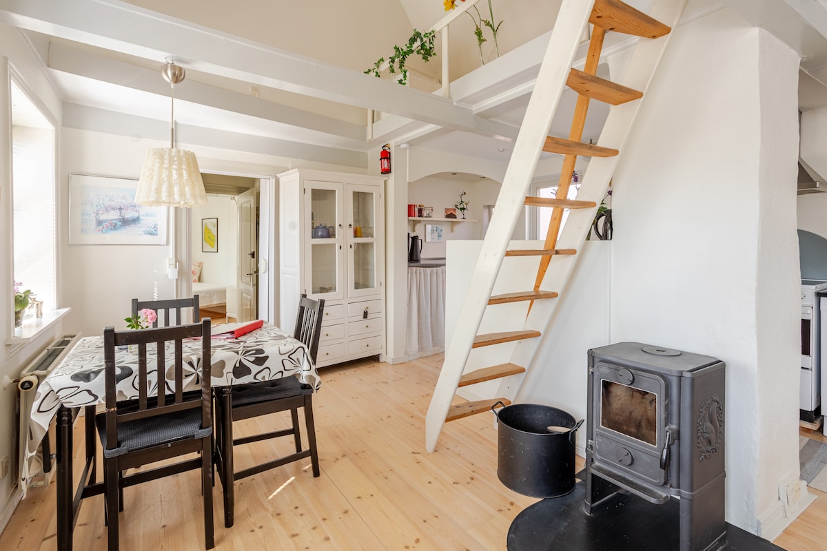 A dining area is furnished with a table and four chairs, set against wooden flooring. A staircase leads to an upper level. A wood stove is positioned nearby, while a pendant light hangs from the ceiling, adding a touch of coziness to the space.