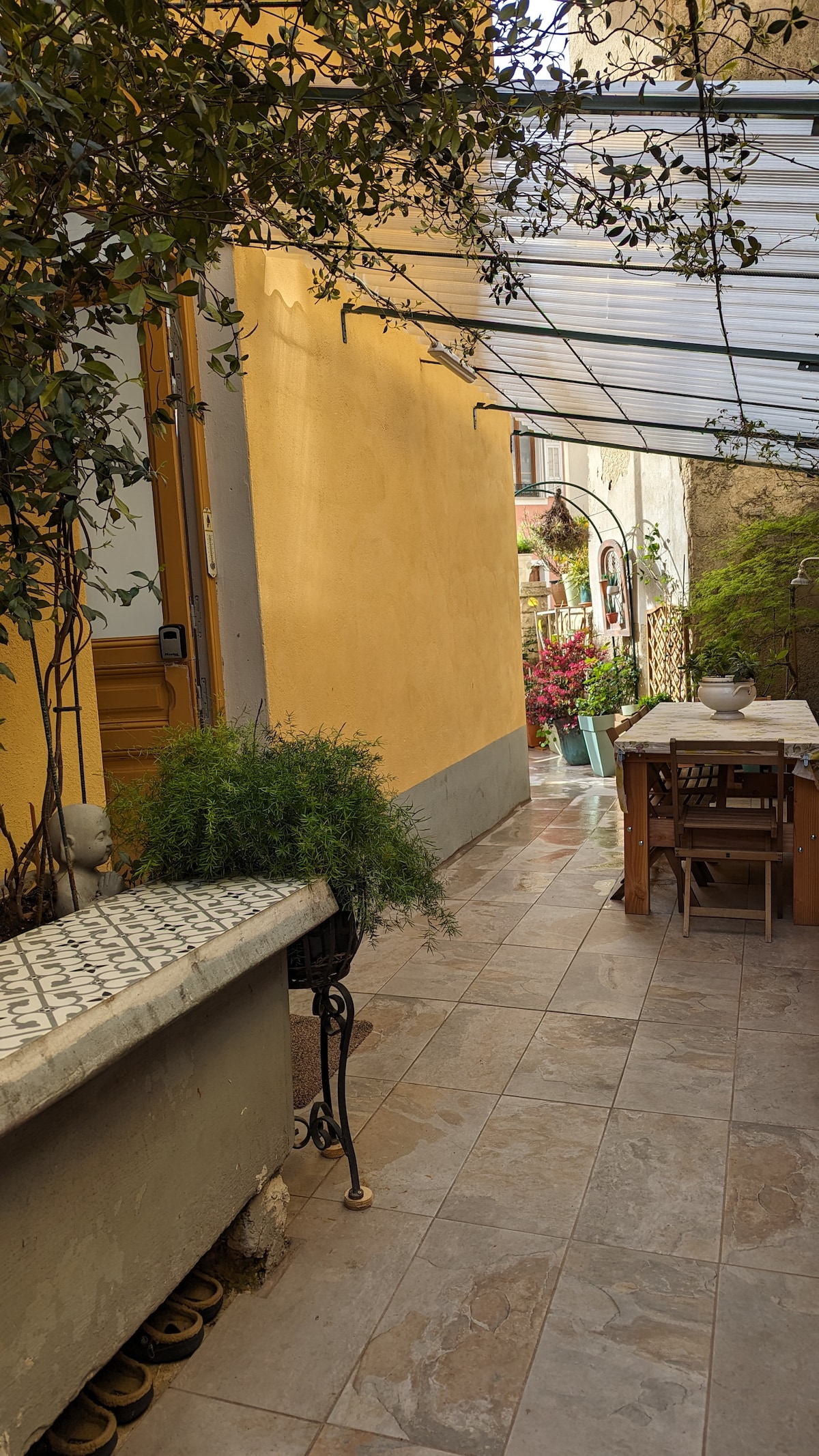 A sunlit terrace area is lined with a smooth tiled floor, bordered by a vibrant yellow wall. A wooden table is centrally placed, accompanied by four chairs. Lush greenery is visible in pots along the wall, leading towards a scenic view of blooming plants.