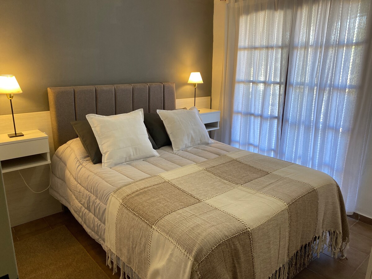 A comfortable sleeping area features a spacious bed with a neutral-toned, textured blanket. Soft pillows are arranged at the head of the bed. Two bedside lamps provide gentle lighting, and large windows allow natural light in, creating a serene atmosphere.