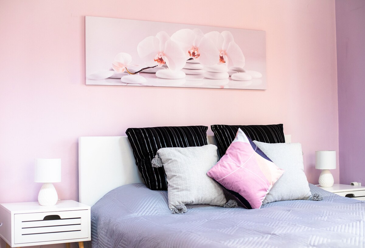 A bedroom setup features a spacious bed with decorative pillows and a soft gray blanket. A large floral artwork in gentle hues adorns the wall above the bed. Bedside tables with lamps flank either side, contributing to a pleasant and restful environment.