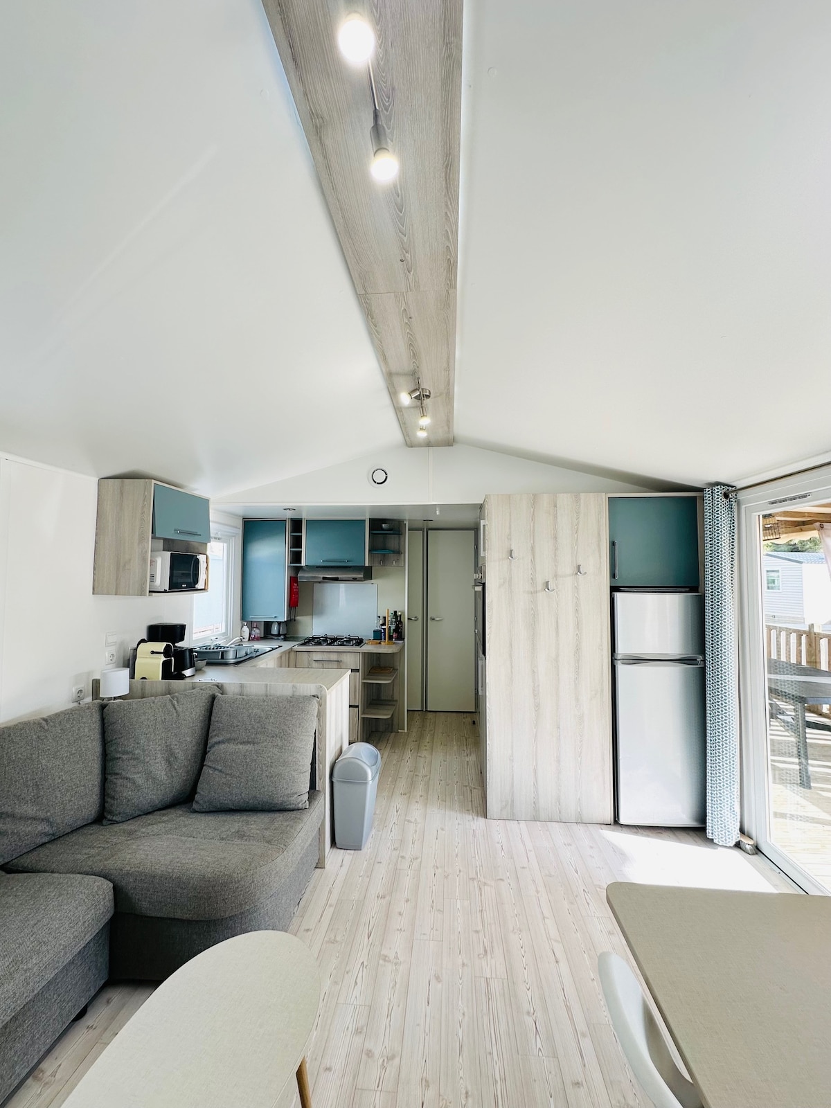 The interior space showcases a modern, open-plan living area with soft seating and a light wood floor. A fully equipped kitchen features sleek cabinets and appliances while natural light floods in through the patio doors, highlighting the comfortable layout and functional design.