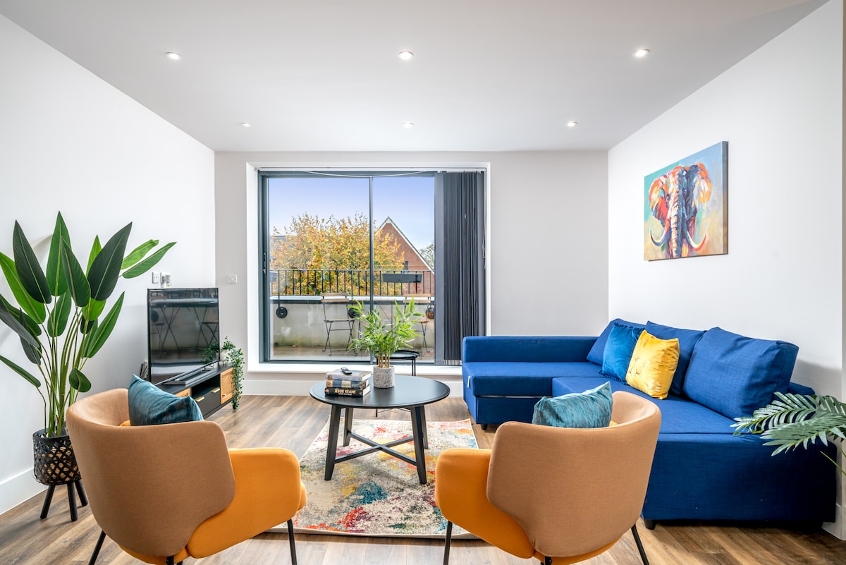 A bright living area features large glass doors that open to a balcony. A blue sectional sofa and two cozy armchairs are arranged around a circular coffee table. A colorful rug adds warmth, while potted plants bring a touch of nature to the space.