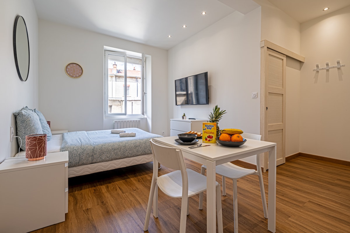 A bright studio features a spacious bed adorned with light bedding, complemented by a television mounted on the wall. A small dining table with white chairs stands beside a window, which provides natural light and a view of the outdoors.