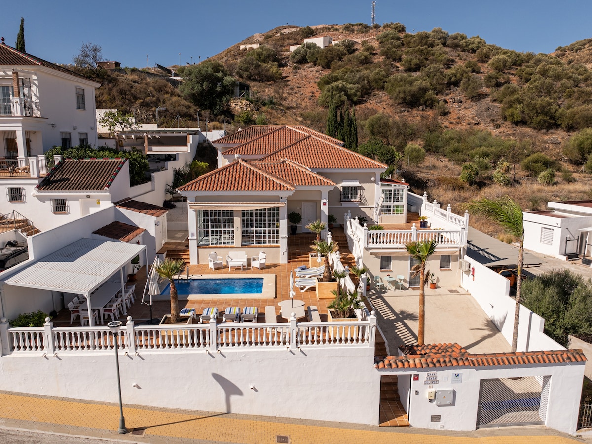 An aerial view of Villa Casa Vista del Rey showcases a spacious outdoor area with a private pool surrounded by sun loungers. Palm trees and garden furniture create a relaxed environment. The villa features white walls and terracotta roofing, set against a backdrop of rolling hills.