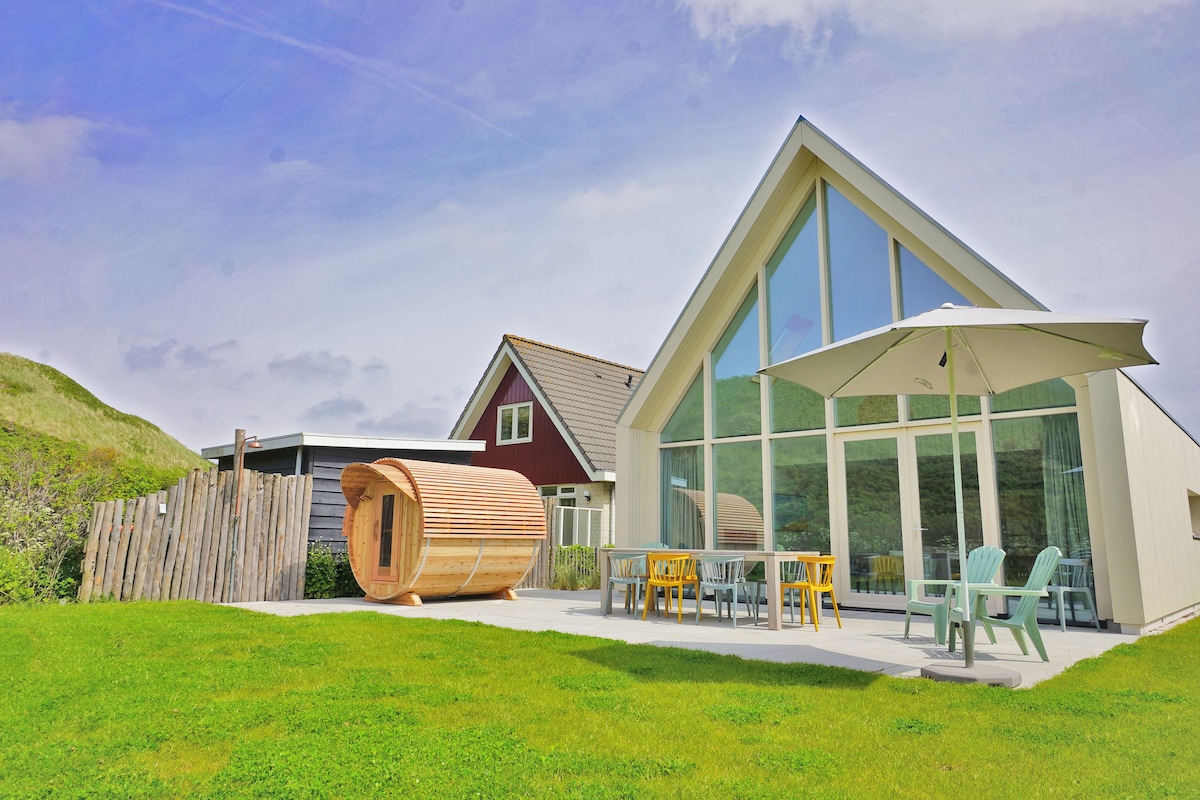 The exterior of Villa Lepelaar features a modern, triangular-roofed structure with large glass windows. A spacious patio is furnished with seating, including pastel-colored chairs and a table. A wooden sauna is visible to the side, surrounded by lush green grass.