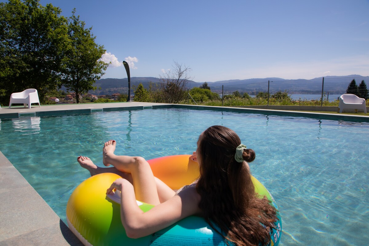 A serene pool area is captured, featuring clear turquoise water surrounded by a stone patio. A colorful inflatable float is positioned in the water. In the background, lush green hills and a clear blue sky provide a scenic view, enhancing the tranquil outdoor atmosphere.