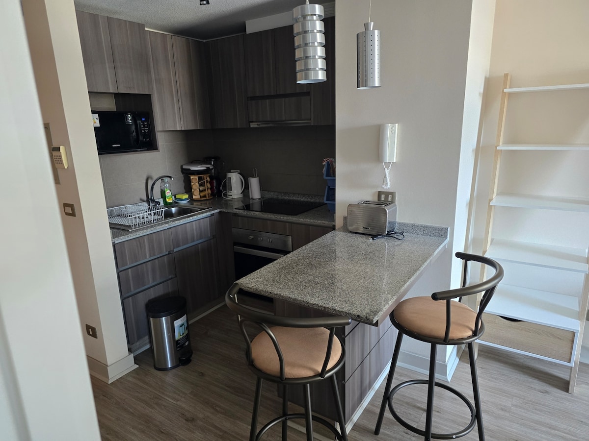 The kitchen features modern cabinetry with a granite countertop, equipped with essential appliances. A small table with two stools is positioned for dining. Natural light illuminates the space through a nearby window, enhancing the overall functionality of the area.