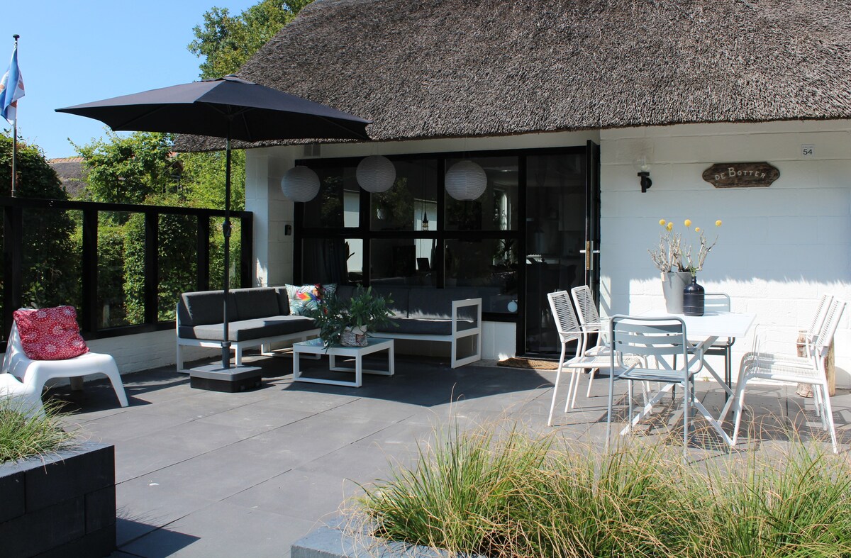 A spacious terrace features a modern lounge area with a sectional couch and a table. A large umbrella provides shade, while the surrounding greenery adds privacy. A dining table with chairs is visible, offering an inviting space for outdoor meals.