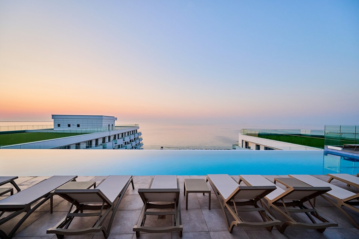 An infinity pool is showcased with multiple sun loungers arranged along the edge. The tranquil sea is visible beyond the pool, blending with the sky at dusk, creating a serene atmosphere. The modern buildings of the resort are visible in the background.