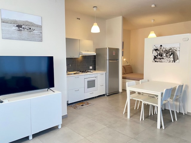 The open-plan living area features modern furniture, including a white table with six chairs and a comfortable sofa bed. A large TV is positioned in front of the dining area, while a well-equipped kitchen showcases sleek cabinets and appliances, including a refrigerator and oven.