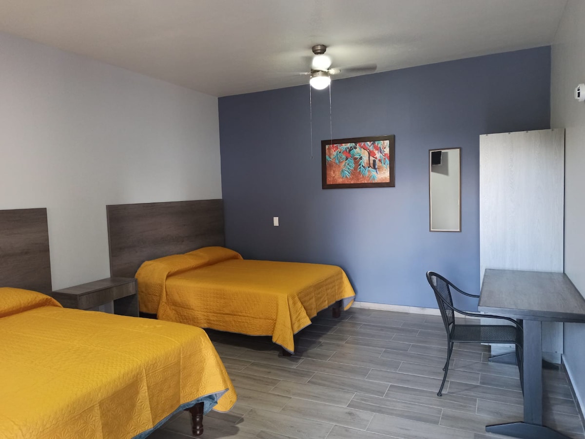 A well-lit guest room featuring two spacious beds with golden bedspreads. A small table and a black chair are positioned against the wall, accompanied by a mirror. Artwork adds a pop of color to the room, which has light wooden flooring.