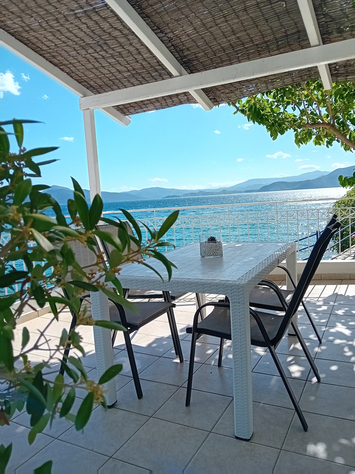 A shaded outdoor dining area is presented with a white table and four black chairs. Lush green foliage frames the scene, with a serene view of the sea and mountains in the background. The space is bathed in natural light, enhancing the tranquil atmosphere.