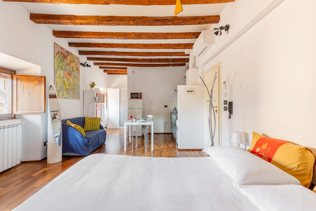 The studio features exposed wooden beams and a spacious layout. A large bed is positioned prominently, accompanied by a sofa in the seating area. A dining table is present, with a kitchenette visible in the background, including modern appliances and ample natural light from the windows.