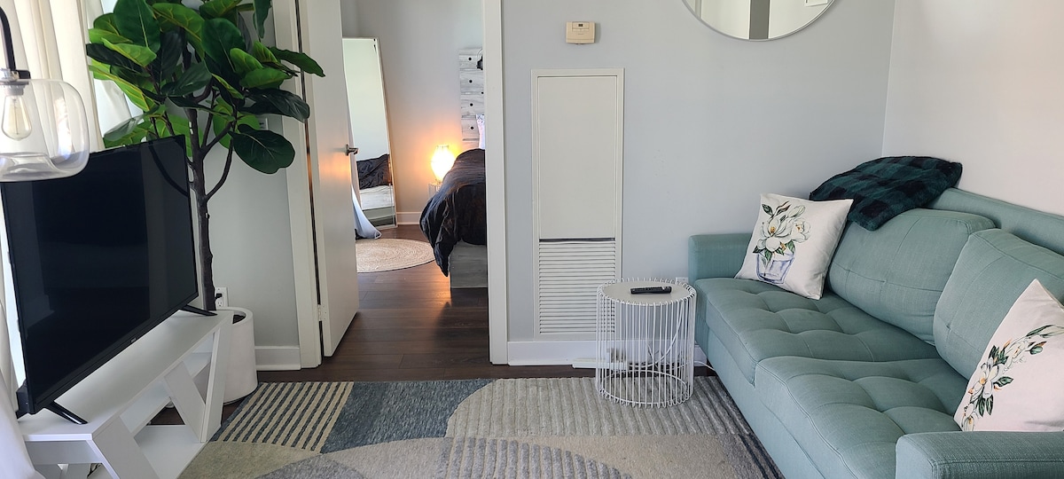A cozy living area is furnished with a light blue sofa adorned with decorative pillows. A small white rug with geometric patterns covers the floor. Adjacent to the space, a doorway opens to a bedroom, illuminating the room with soft light. A tall indoor plant adds a touch of greenery.
