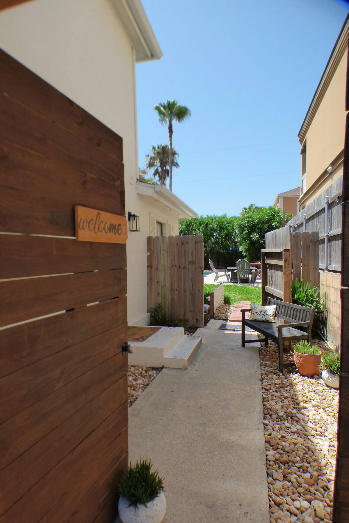A pathway leads through a wooden gate, featuring a welcome sign. Flanking the path are potted plants and decorative stones, with a small seating area set against the privacy fence. Lush greenery is visible at the end of the pathway, under a clear blue sky.