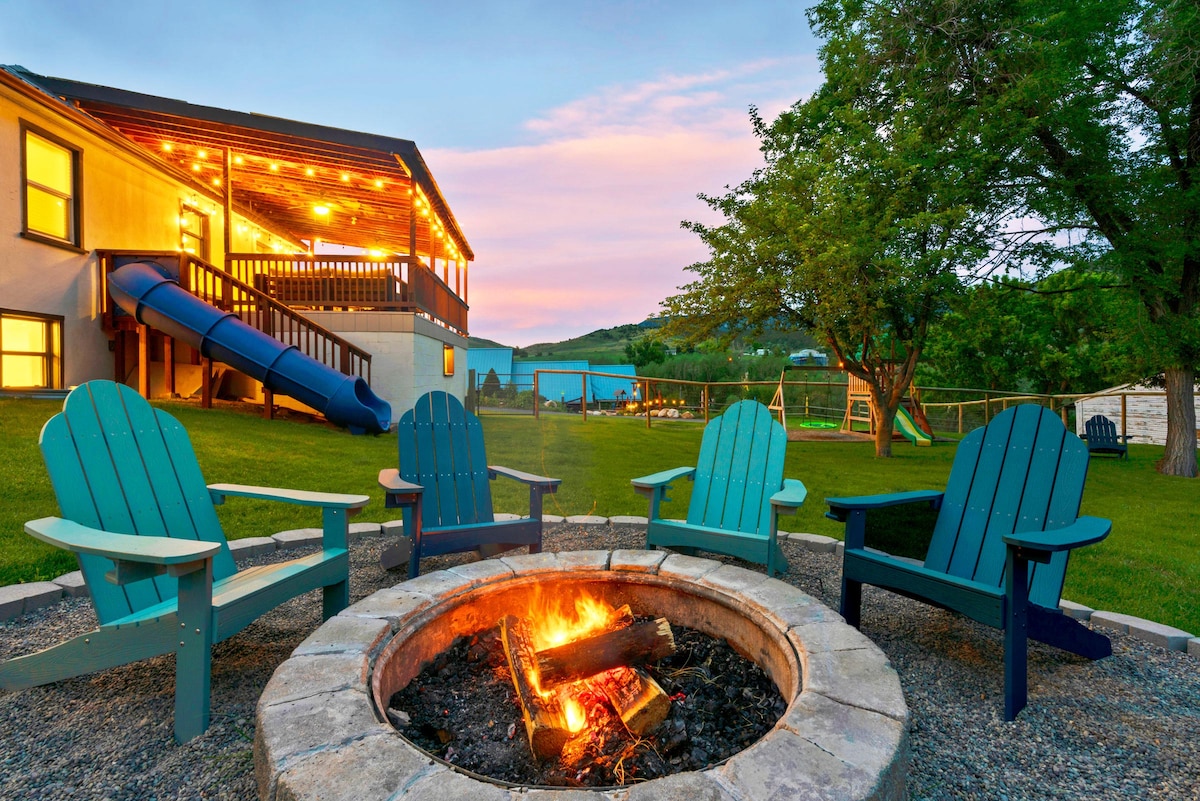 A fire pit surrounded by four blue Adirondack chairs is positioned in the large yard. In the background, a wooden elevated patio is visible, complete with a custom tube slide leading down to the grass. Soft lighting reflects a warm ambiance against the evening sky.