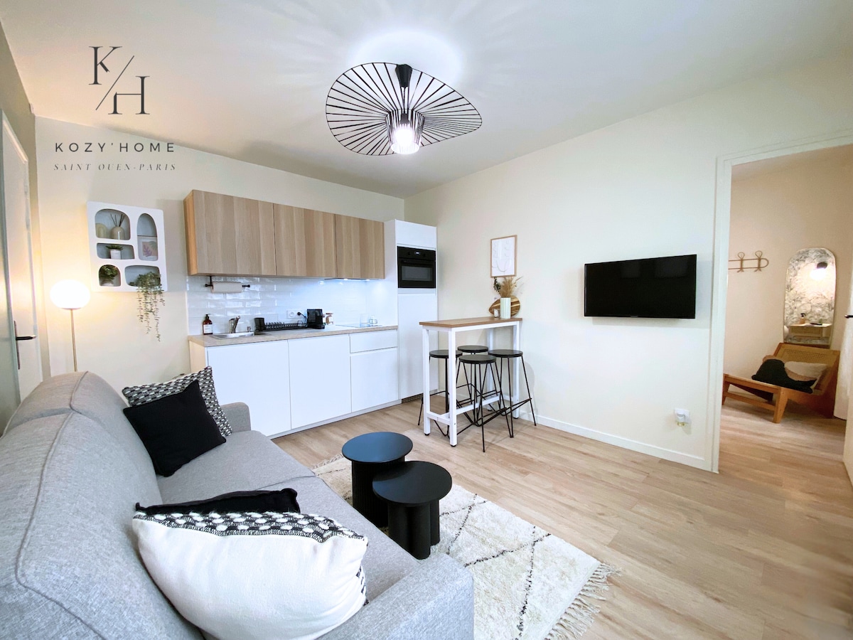 A modern living space is showcased, featuring a comfortable gray sofa adorned with decorative cushions. A compact kitchen with white cabinetry and appliances is present in the background. A dining area with high stools provides additional seating, while a smart TV is mounted on the wall.