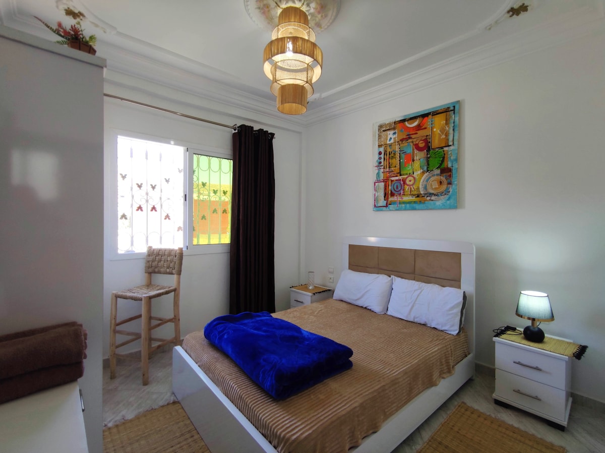 The bedroom features a spacious bed with crisp white linens and a dark blue blanket. A colorful piece of wall art adds a vibrant touch. Natural light streams through a window, complemented by dark curtains. A bedside lamp and a wooden stool are present in the room.