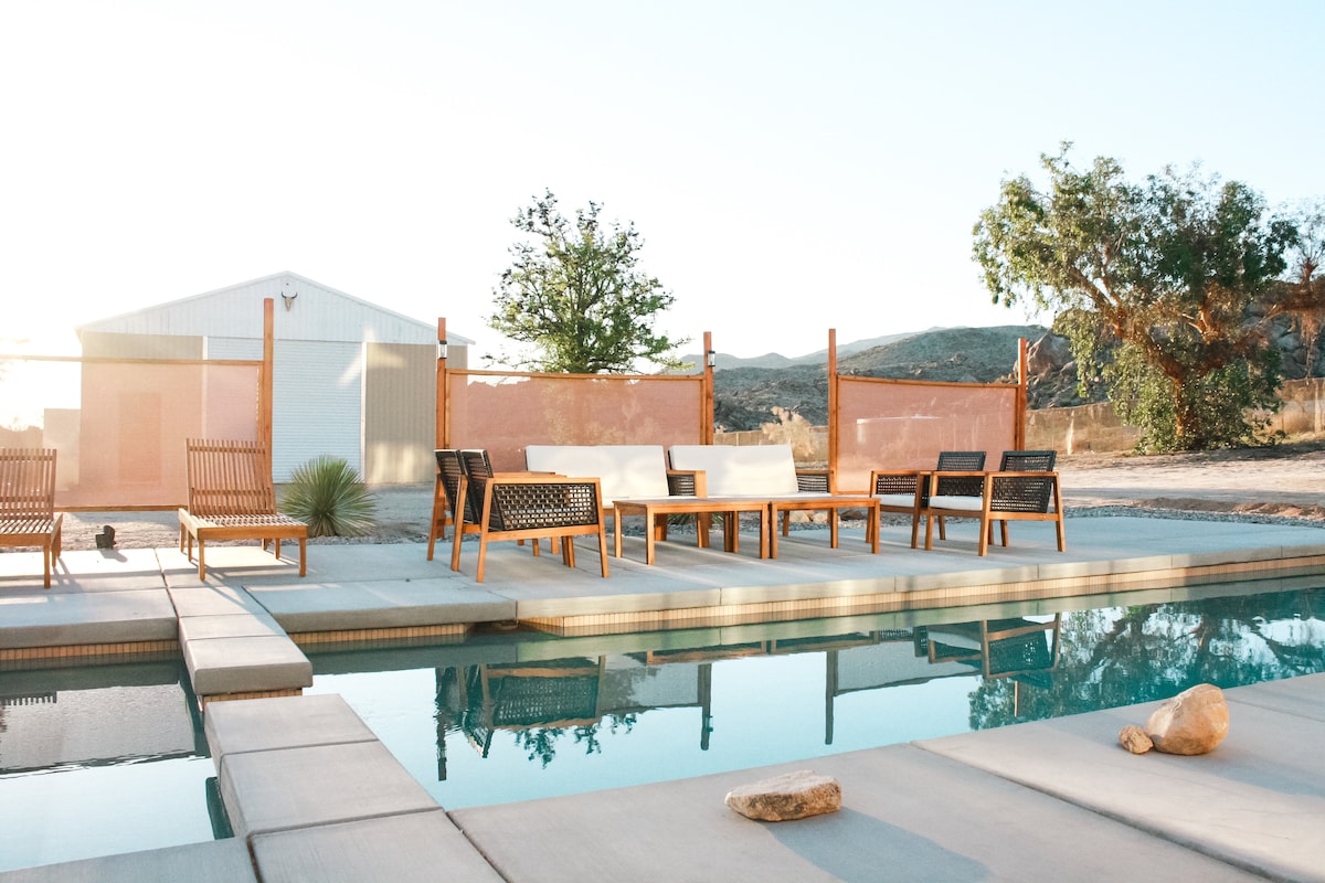 A serene outdoor area is highlighted by a saltwater pool, reflecting the clear sky. Wooden furniture with woven details is arranged thoughtfully around the pool, providing ample seating. A barn-like structure and desert vegetation are visible in the background, enhancing the tranquil atmosphere.